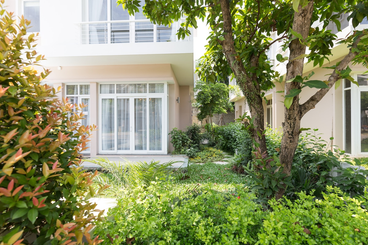Lush greenery surrounds the villa, with vibrant plants including small shrubs and trees. Sunlight filters through the leaves, gently illuminating the outdoor space. The villa's large glass windows reflect the garden, creating a harmonious connection between the indoor and outdoor areas.