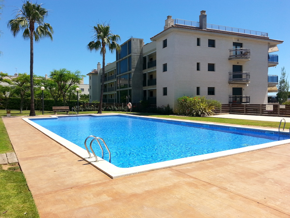 A spacious outdoor pool is surrounded by well-kept gardens and palm trees, inviting relaxation. The pool area features a light blue water surface and has steps leading into it. Nearby buildings, included in the image, display a smooth, modern design.