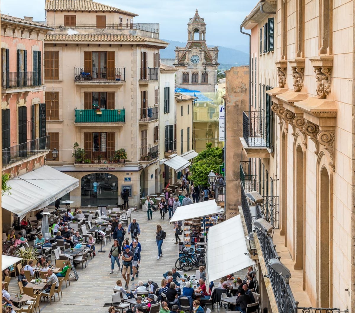 A lively street scene showcases a bustling plaza filled with people enjoying outdoor seating at cafés. Surrounding buildings possess traditional architecture, with balconies overlooking the square. The backdrop includes a clock tower and distant mountains under a partly cloudy sky.