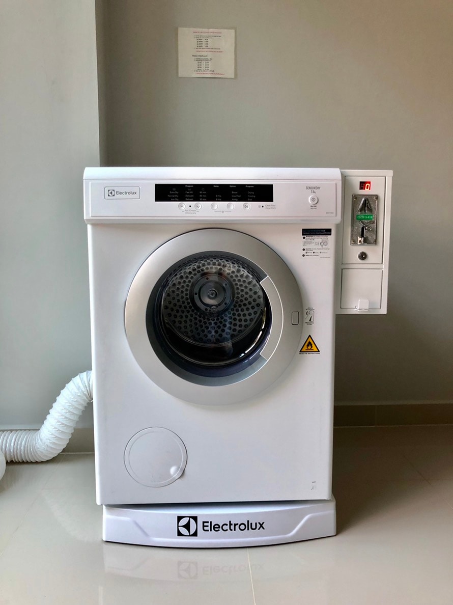 A modern washing machine is positioned against a smooth wall. The machine's front is white with clear control buttons and a circular door. A hose extends from the back, and a power outlet is visible above it, ensuring easy access for operation.