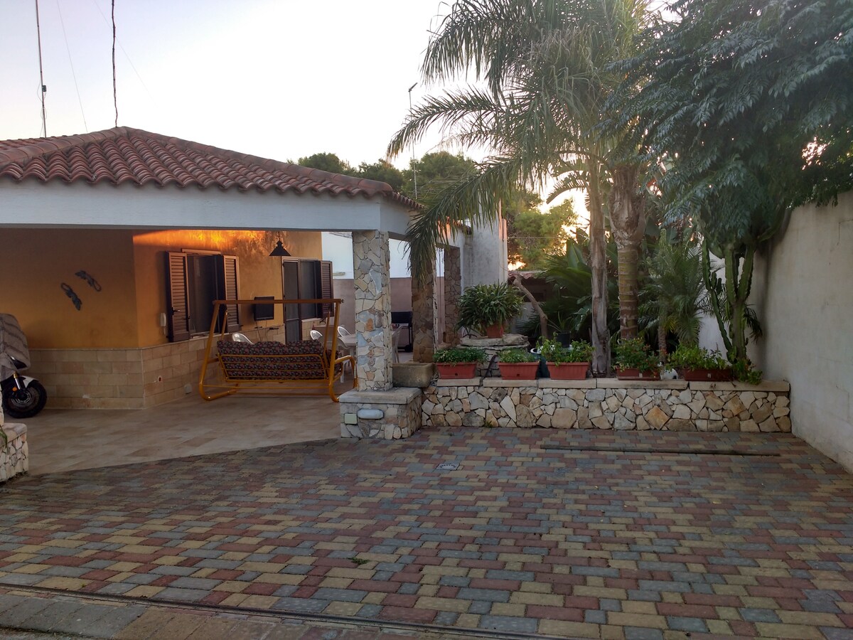 An outdoor area features a stone-paved driveway leading to a charming house. A swinging bench is placed in the shaded porch, surrounded by lush green plants and flower beds. The warm glow of sunset highlights the inviting atmosphere of the space.