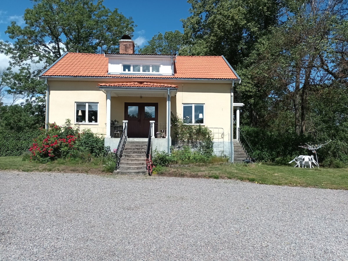 A two-story house is depicted with a yellow exterior and a red-tiled roof. Steps lead up to the front door, flanked by two windows on each side. Surrounding greenery and flowerbeds add natural beauty to the setting, while a gravel area provides ample parking space.