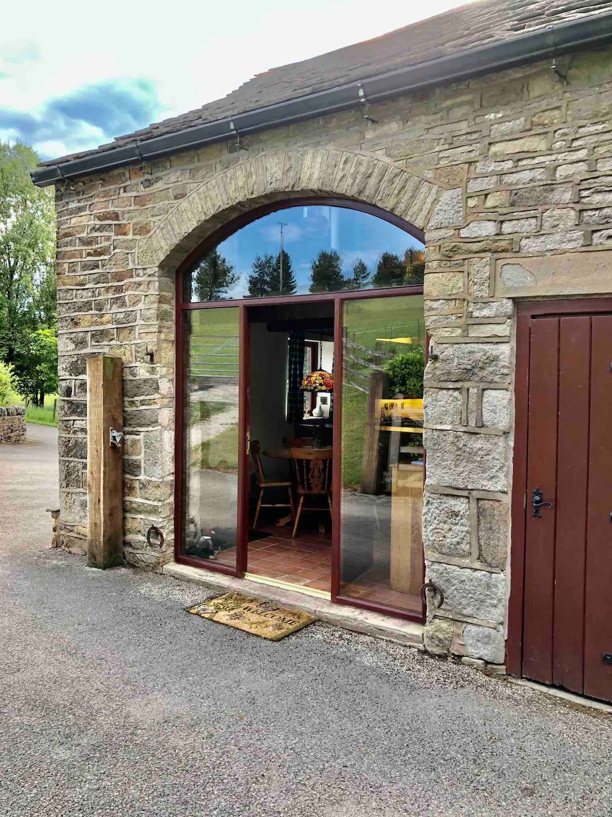The entrance is framed by a large arched window, allowing natural light to fill the space. Stone walls accentuate the rustic charm of the converted barn. A wooden door is visible to the right, leading into the interior.