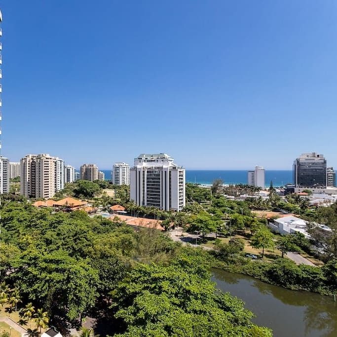 A panoramic view from the 14th floor reveals lush greenery alongside a tranquil lagoon, framed by modern buildings. The blue ocean is visible in the distance, with clear skies above. The urban landscape showcases a harmonious blend of nature and city life.