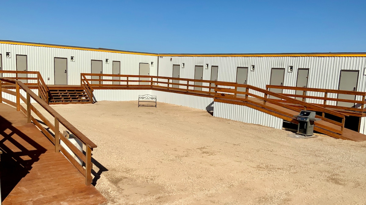 An outdoor space features a circular arrangement of individual rooms with access via wooden walkways. Benches are positioned centrally, while outdoor lighting is visible near each door. The surrounding area is covered with gravel, providing a low-maintenance environment.
