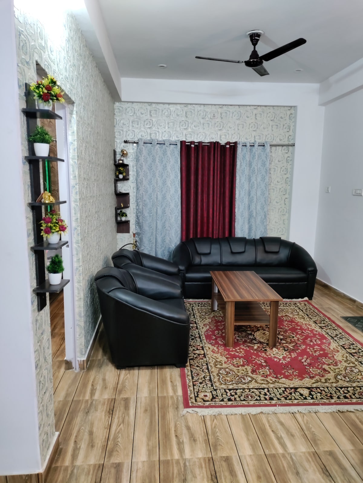 A living area features a sleek black sofa set, consisting of a three-seater and two armchairs, arranged around a wooden coffee table. A decorative red and gold area rug lies on the floor. Light blue curtains frame a window, complementing the patterned wall.