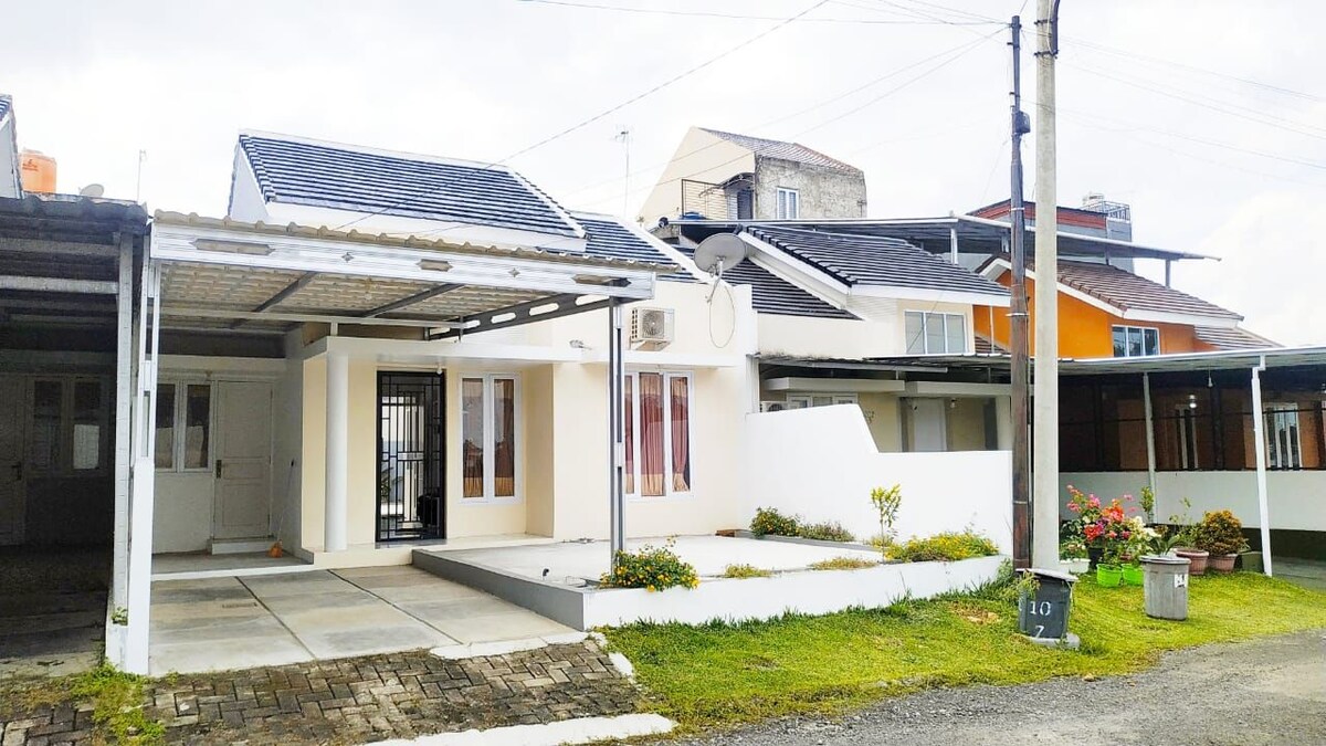 The front exterior of the house features a clean and modern design with an accessible entrance. A spacious paved driveway leads to the entrance, adorned with small garden beds that add greenery. Large windows provide natural light to the interior while enhancing the home’s inviting appearance.