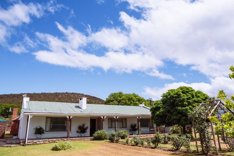 The exterior of the lodge is shown, featuring a single-story structure with a green roof and a white facade. Lush greenery surrounds the building, and a trellis with climbing plants adds to the inviting garden space. The background includes rolling hills under a partly cloudy sky.