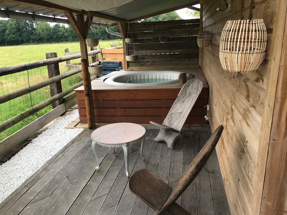A wooden terrace features a relaxing jacuzzi surrounded by a rustic design. A light-colored coffee table sits alongside a sculptural wooden chair, while a natural setting with greenery and a wooden fence is visible in the background.