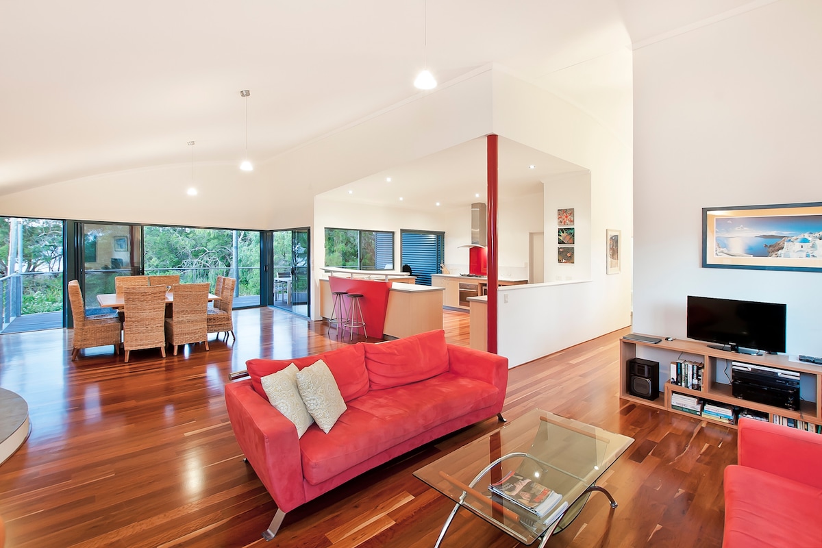 An open plan living area is depicted with polished hardwood floors, featuring comfortable red couches and a glass coffee table. A dining area with a large table and woven chairs is visible alongside the modern kitchen. Expansive windows allow natural light and views of the surrounding greenery.
