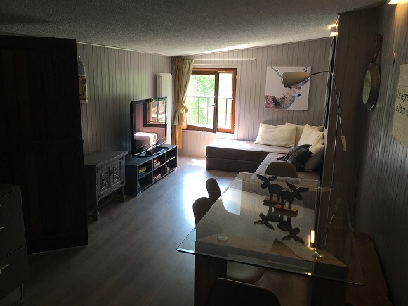 A cozy living area is depicted, featuring a large wall-mounted television opposite a sectional sofa. A dining table with six chairs sits adjacent. Natural light filters through the window adorned with a curtain, enhancing the wood-paneled walls and light-colored flooring.