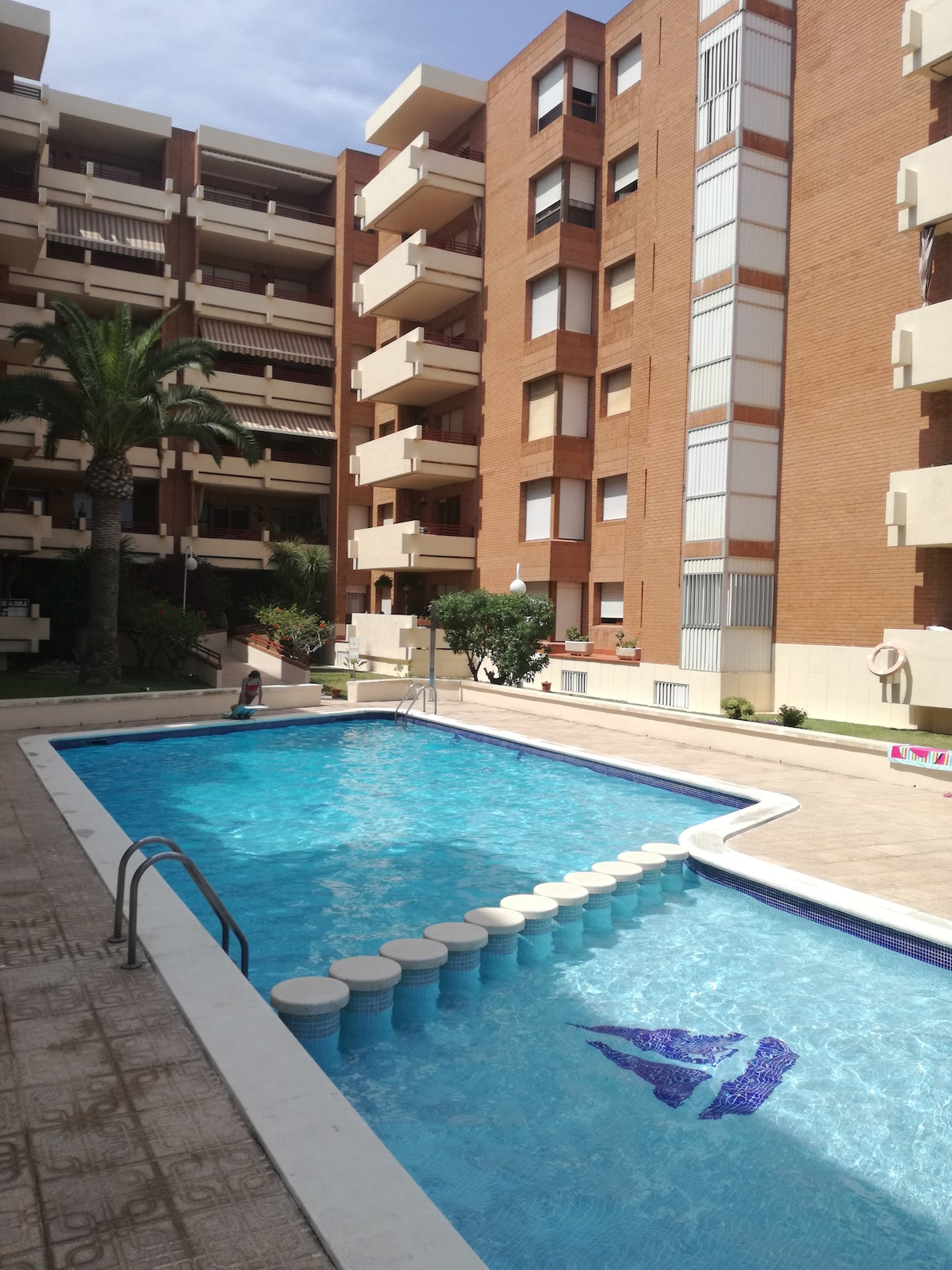 A communal swimming pool is featured in the foreground, surrounded by a spacious patio. Sun loungers are positioned along the edge. The building's exterior is visible in the background, showcasing several balconies and greenery that enhances the overall setting.