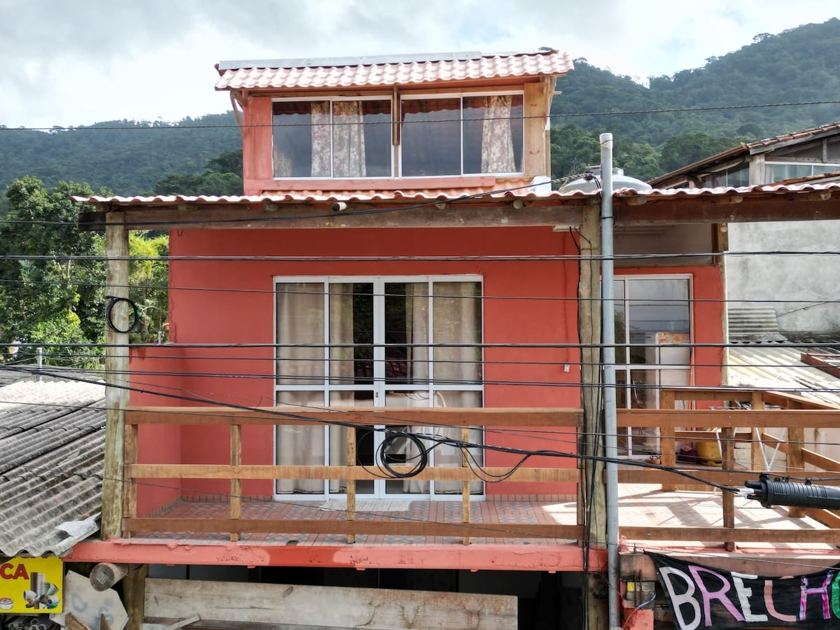 An exterior view of the sturdy red chalet displays a welcoming balcony with wooden railings. Large glass doors are framed by curtains, allowing for ample natural light. The surrounding landscape features rolling hills in the background, contributing to a relaxing atmosphere.