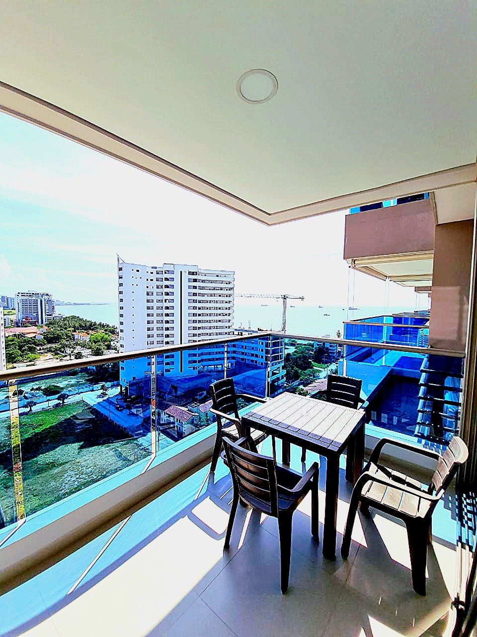 A private balcony features a dining setup with a small table and four chairs, surrounded by glass railings. The view encompasses a distant horizon, the ocean, and the vibrant surroundings of the condominium, highlighting a blend of city and nature.
