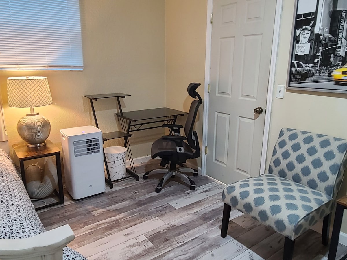 A functional workspace is presented with a desk and ergonomic chair. A lamp provides soft lighting beside a stylish accent chair. An air conditioning unit is positioned nearby, and a door leads to the adjoining area. Textured flooring adds depth to the room.