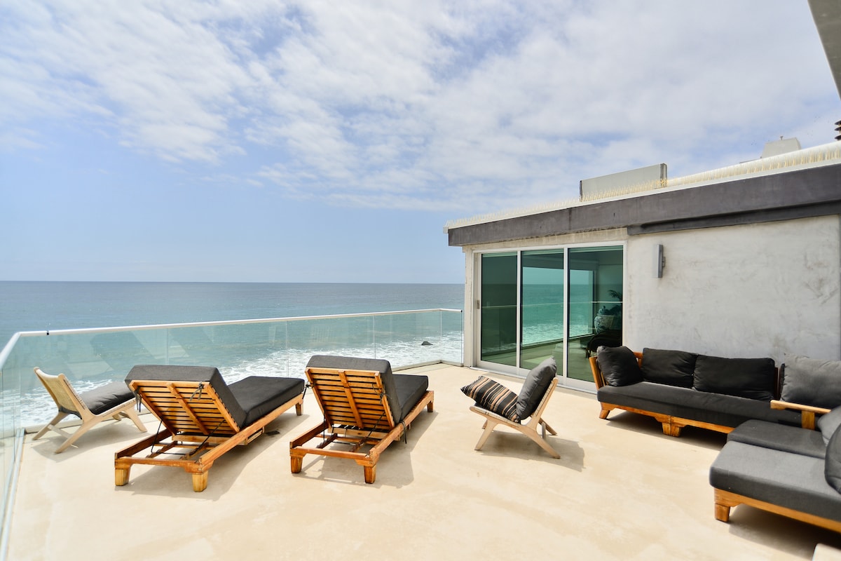 A spacious balcony overlooks the ocean, featuring lounge chairs and a sectional sofa. Glass railings provide unobstructed views of the water, while the concrete surface enhances the modern aesthetic. The soothing sound of waves is part of the serene atmosphere.