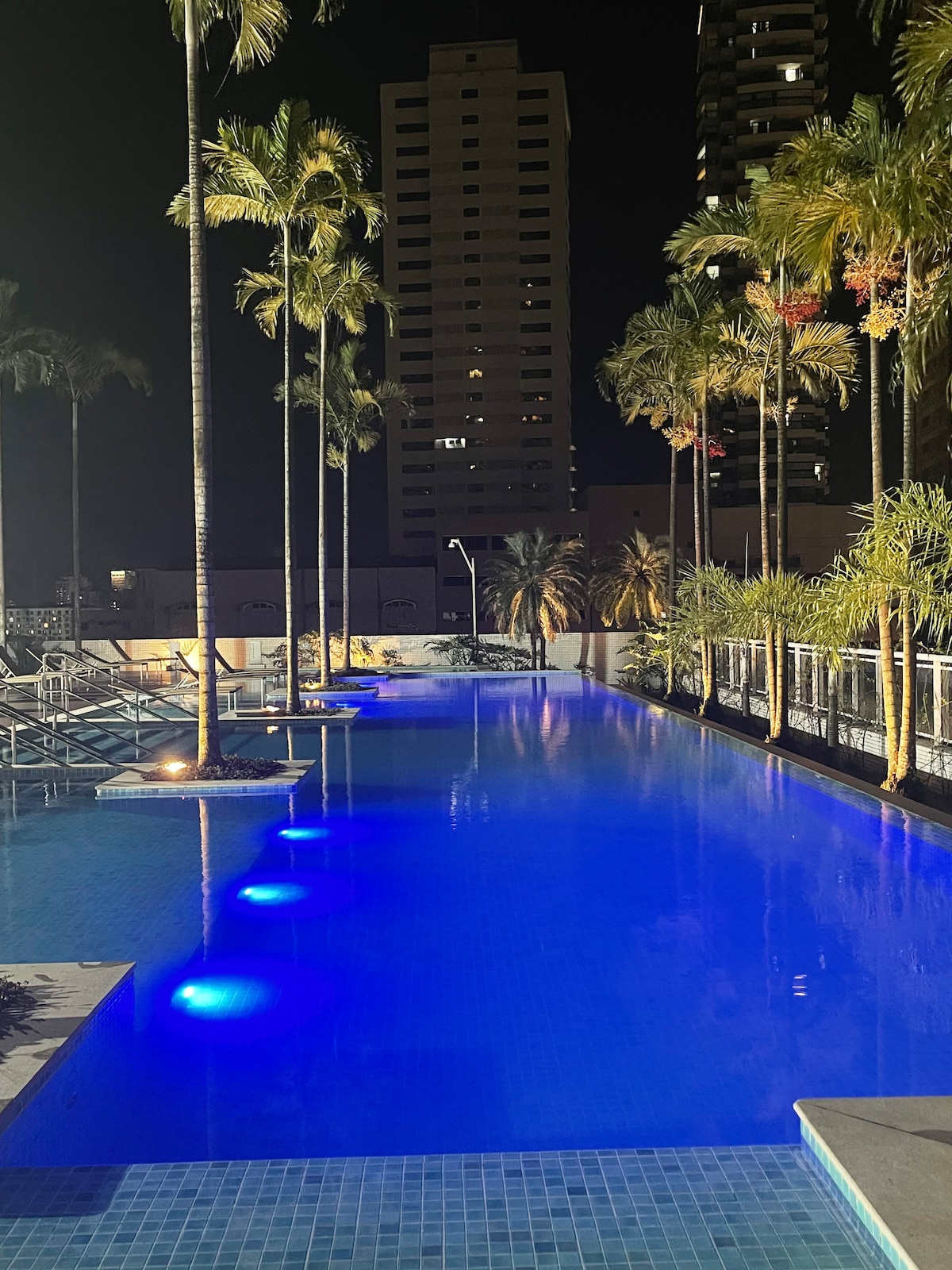 A tranquil pool is illuminated by soft blue lights at night, surrounded by palm trees and modern high-rise buildings. Lounging chairs are positioned along the poolside, creating a serene ambiance for relaxation.