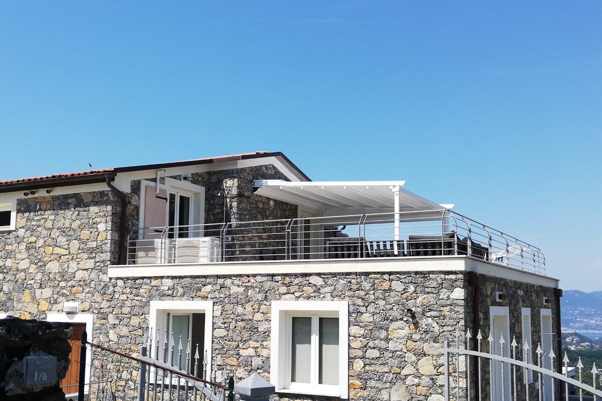 The exterior of the stone villa is highlighted by a spacious terrace featuring protective awning cover. Large windows allow for ample natural light. The building is surrounded by a gated fence, with a clear blue sky in the background, enhancing the serene setting.