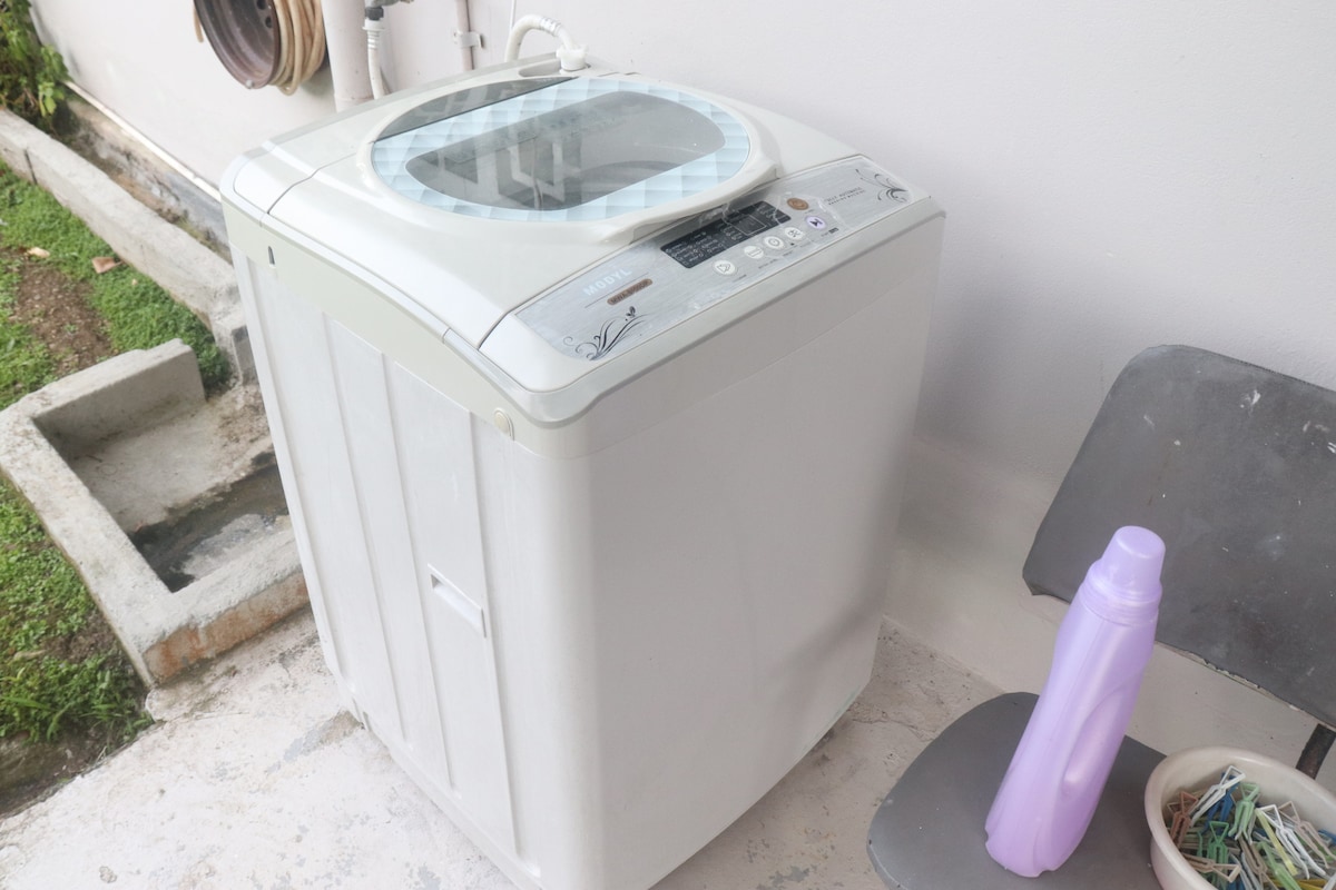 A washing machine is located outside the flat, positioned on a concrete surface beside a chair. A water bottle is placed next to the chair, providing a convenient space for guests. A grassy area is visible in the background, suggesting a clean environment.