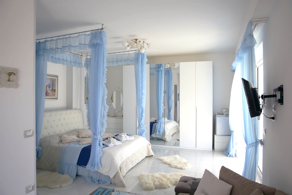 The spacious bedroom features a large, upholstered bed draped with sheer blue fabric. A light-colored quilt and throw pillows adorn the bed. A mirror and wardrobe are visible, reflecting natural light. Fluffy rugs are placed on the tiled floor, enhancing the cozy atmosphere.