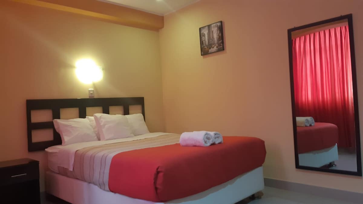 A well-appointed room features a spacious bed with white linens and a red accent blanket. A large mirror reflects the interior, and soft lighting highlights the warm walls. Red curtains frame a window, allowing natural light to enhance the cozy atmosphere.