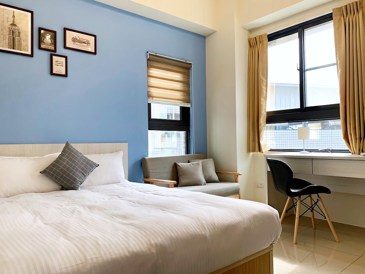 A bright sleeping area features a spacious bed with crisp linens, complemented by a decorative pillow. A cozy armchair sits beside a study desk, and sunlight filters through a large window adorned with light curtains, illuminating the soft blue wall.