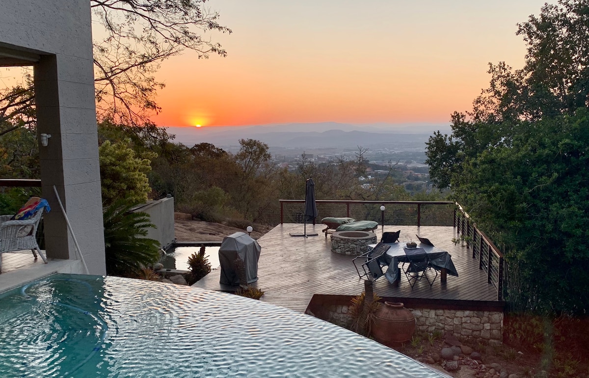 A serene outdoor space features a swimming pool with an infinity edge overlooking a sunset. Lounge chairs and a table for six are visible on the wooden deck, surrounded by trees and distant hills. A grill and a patio umbrella are also present.