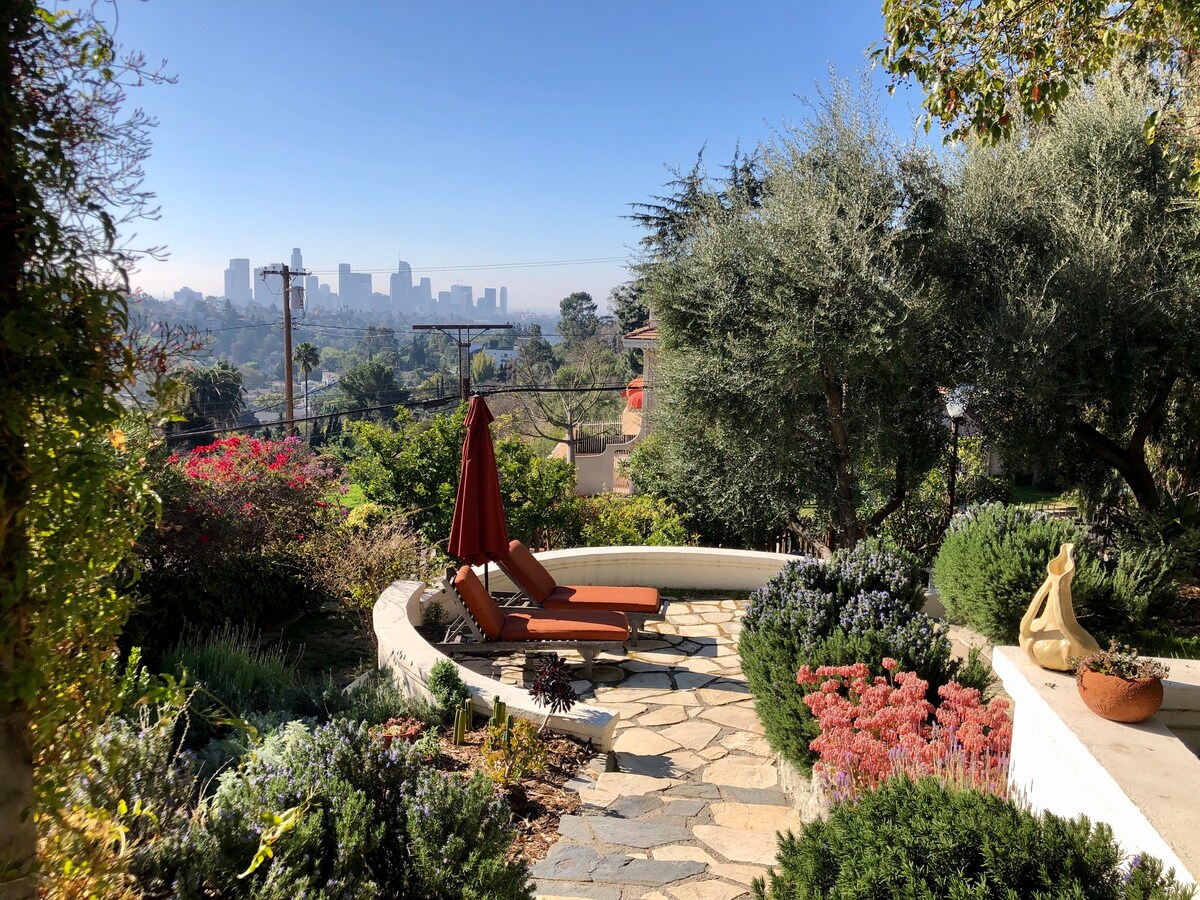 Lounge chairs are positioned on a stone patio, surrounded by vibrant foliage and flowers. An umbrella provides shade, and a view of the city skyline is visible in the distance, framed by lush greenery and blooming plants.