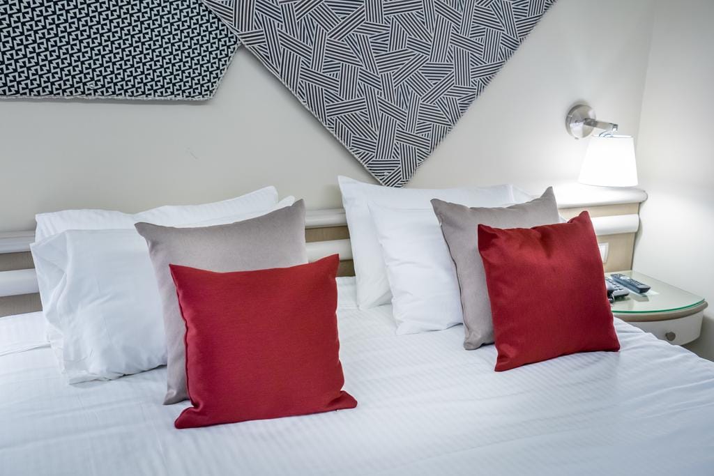 A well-arranged sleeping area features a spacious bed with white linens and decorative pillows in shades of red and beige. Geometric wall art adds a modern touch, while a bedside lamp provides soft illumination.