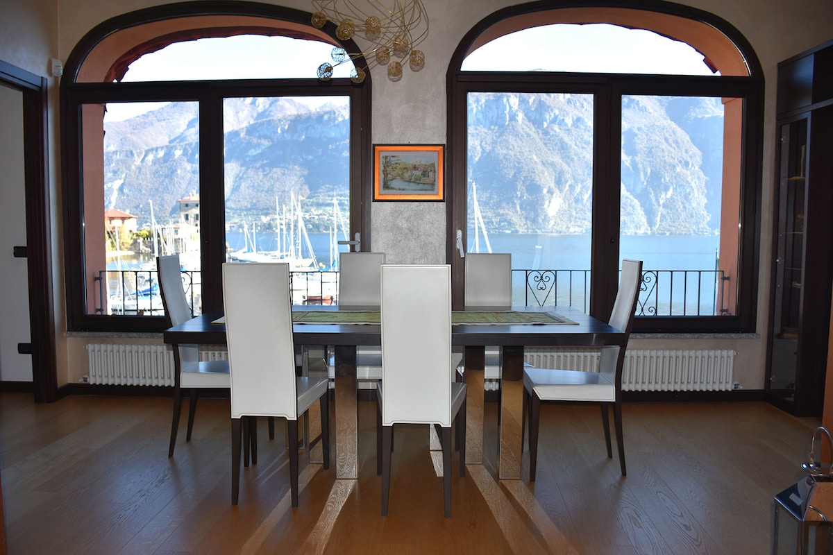 A dining area is highlighted by a long wooden table surrounded by six white chairs. Large arched windows frame a panoramic view of the bay and surrounding mountains, allowing natural light to illuminate the space.