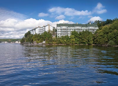 A multi-story building is positioned along the shoreline, surrounded by lush greenery. The reflective surface of the water creates a serene atmosphere, with clouds gently drifting above against a bright blue sky.