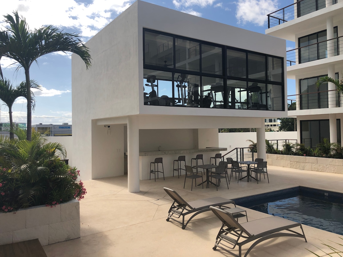 A modern outdoor area features a swimming pool bordered by sun loungers and a spacious terrace. A shaded seating arrangement is positioned near a bar, and a glass-enclosed gym is visible above, showcasing fitness equipment and large windows that invite natural light.