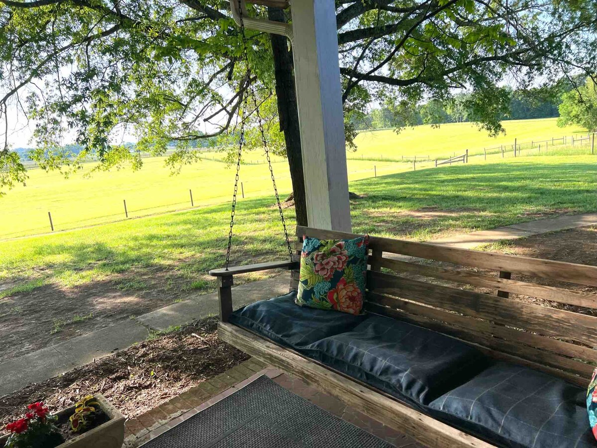 A swing seat with dark cushions is positioned under a large tree, providing shade and comfort. Colorful floral pillows add a touch of warmth. A view of lush green pastures is visible in the background, complemented by a fence line that extends into the distance.
