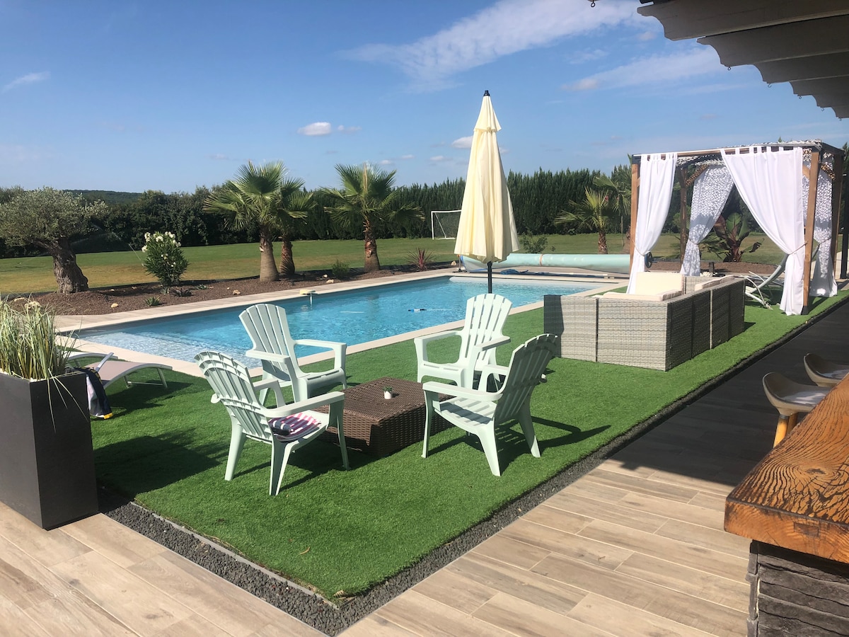 A serene outdoor pool area is surrounded by manicured lawn and palm trees. Comfortable chairs are arranged on the green turf, offering a place to relax in the sun. A shaded seating area with draped fabric and a white umbrella enhances the inviting space.
