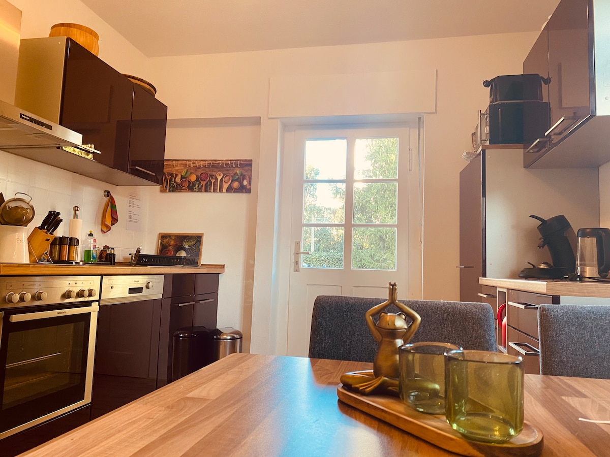 A modern kitchen is presented with dark cabinetry and stainless steel appliances. The dining table, centered in the image, is set with green glass cups, accompanied by a decorative wooden tray. Large windows allow natural light to fill the space, offering views of the garden.
