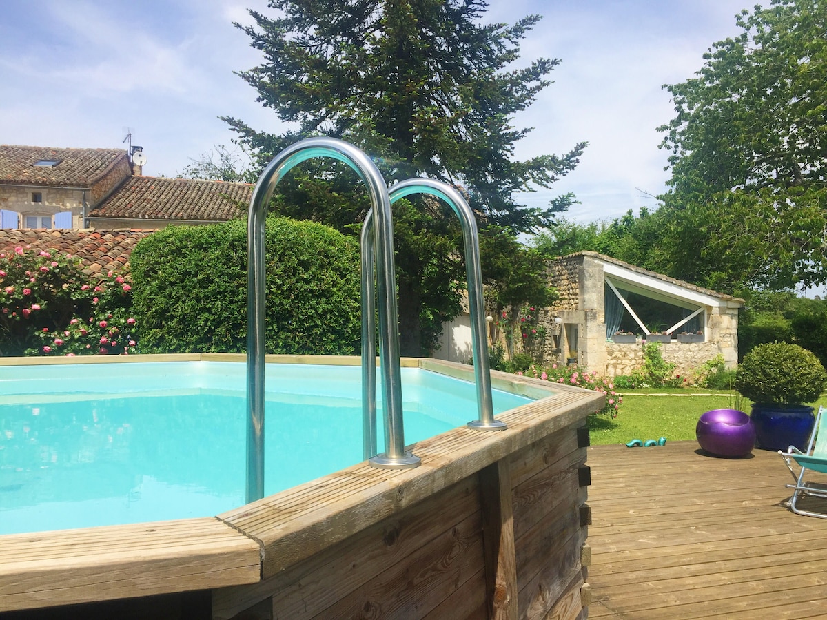 An above-ground pool is framed by a wooden deck, featuring two stainless steel ladder rungs. Lush greenery surrounds the pool area, with flowering shrubs in the background and a charming stone building visible nearby, creating a serene outdoor space.