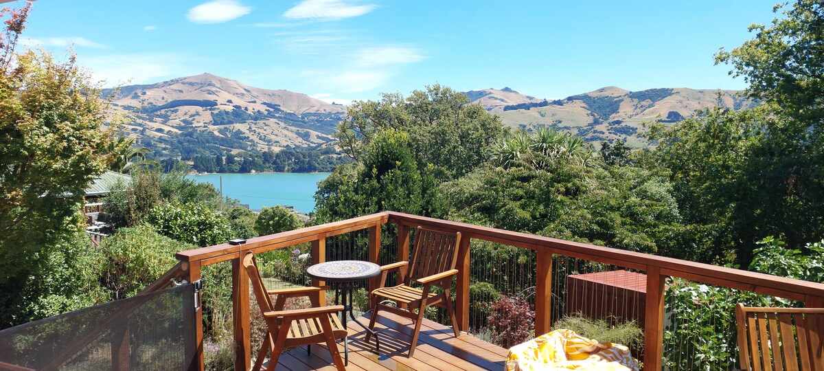 A spacious deck overlooks the scenic Akaroa Harbour, framed by lush greenery and distant hills. Two wooden chairs and a round table are positioned on the deck, inviting relaxation. A blanket is casually draped over the railing, suggesting a cozy outdoor space.