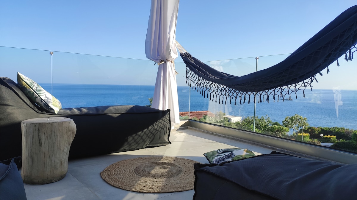 An outdoor seating area is presented with a view of the sea, featuring hammock-style chairs and a wooden stool. A round natural fiber rug is placed on the tiled floor, with soft, draped fabric providing shade against the bright sky.