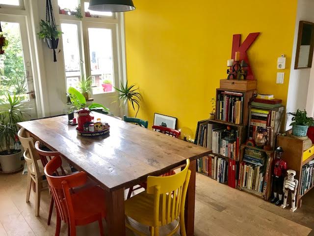 A bright dining area features a large wooden table surrounded by colorful chairs in a variety of hues. A vibrant yellow wall adds warmth, while plants are positioned around the space. A small bookshelf is filled with various items, contributing to the lively atmosphere.