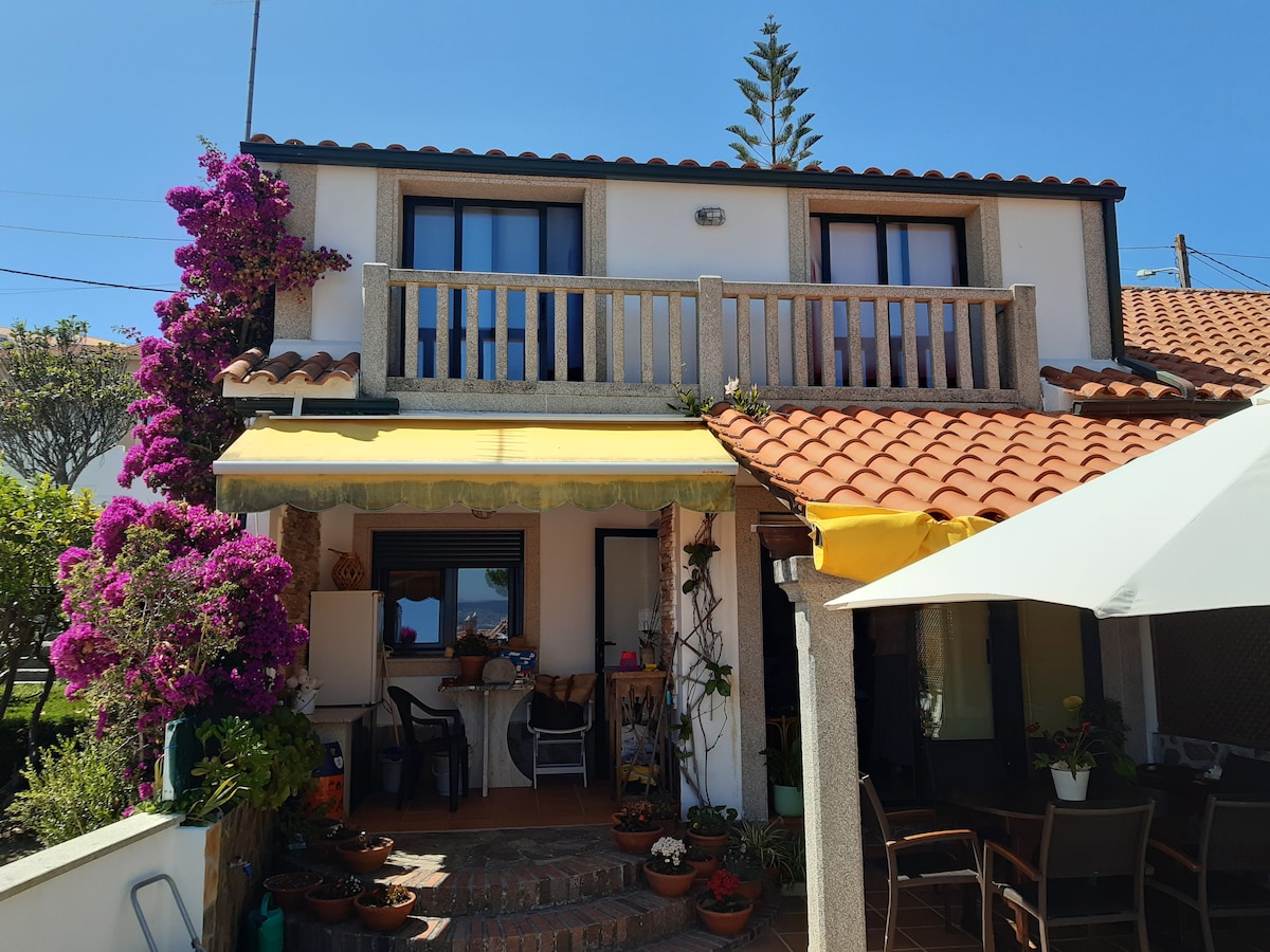 A charming facade of a two-story house is highlighted by vibrant purple flowers climbing its exterior. A balcony extends from the upper level, while a terracotta roof and green awning complement the outdoor seating area with tables and chairs surrounded by potted plants.