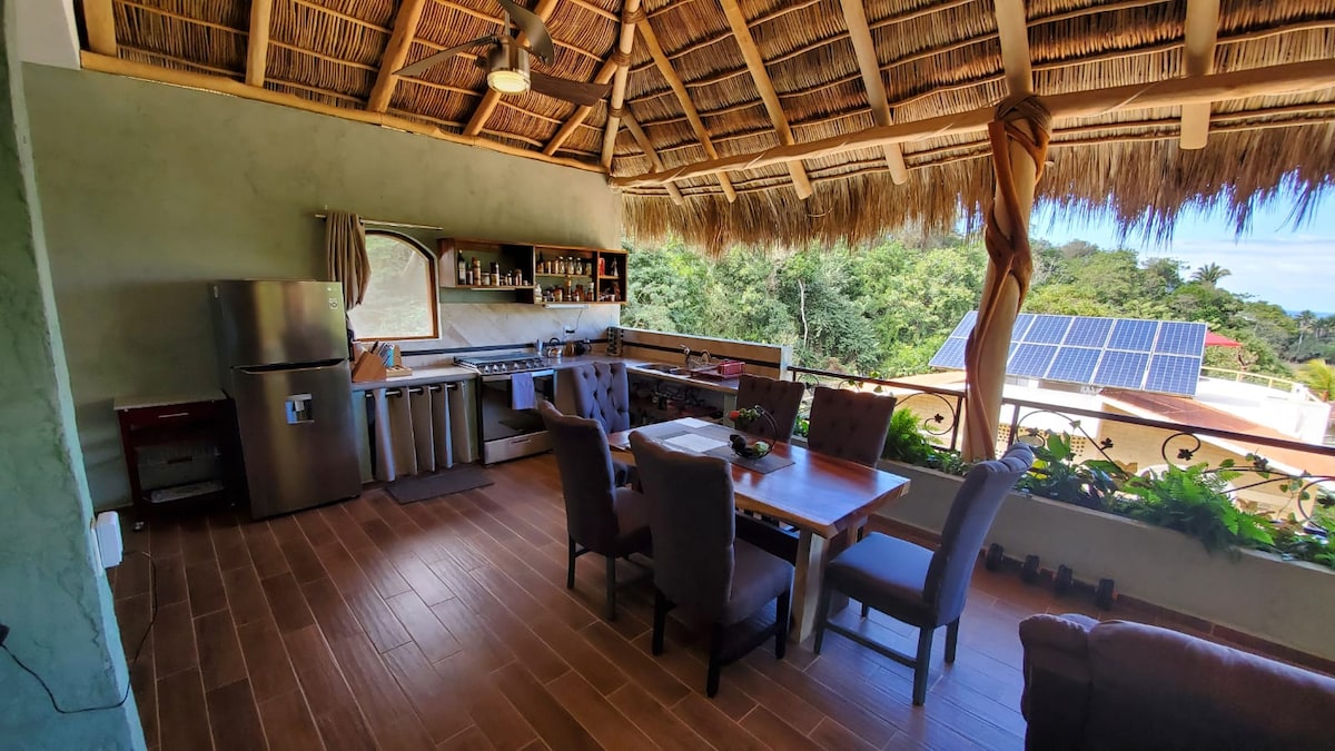 An outdoor kitchen area features a spacious dining table accompanied by chairs, all under a thatched roof. A stainless-steel refrigerator and cooking area are visible, with natural light illuminating the space. Lush greenery is seen through the open wall, enhancing connection with nature.