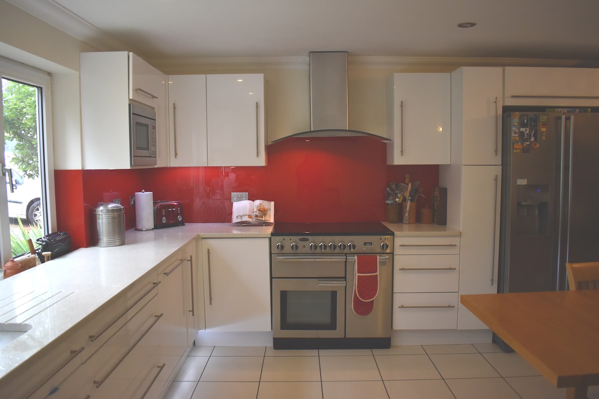 The modern kitchen features white cabinetry paired with a vibrant red backsplash. An induction cooker and stainless steel hood are prominently displayed, complemented by a refrigerator and countertop appliances. Natural light enters through a large window, illuminating the spacious layout.