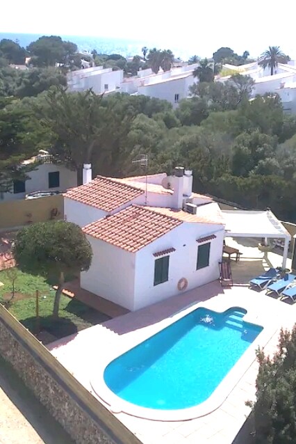 An aerial view of a charming villa set in a tranquil area, surrounded by greenery. A large private pool is visible, along with a terrace featuring sun loungers and a shaded area. The villa's distinct red-tiled roof complements the natural landscape.