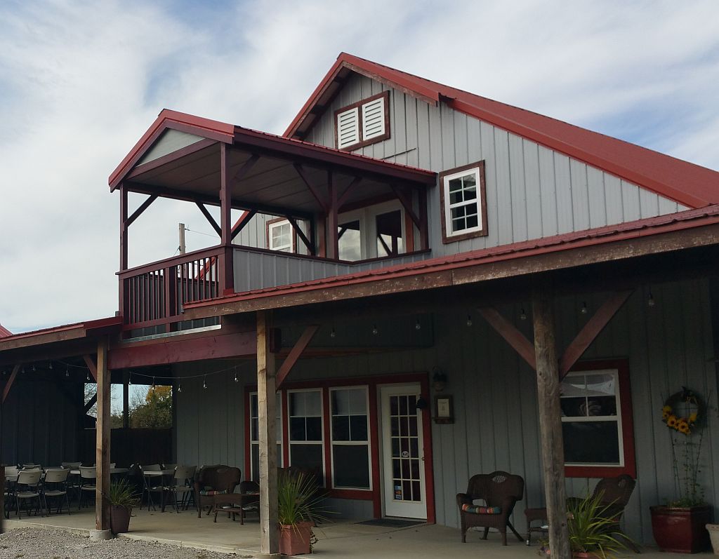 A two-story building features a combination of gray and red siding, with a covered deck on the upper floor. French doors and large windows provide access to the outdoors. The patio area is furnished with seating and decorated with potted plants, creating a welcoming space.
