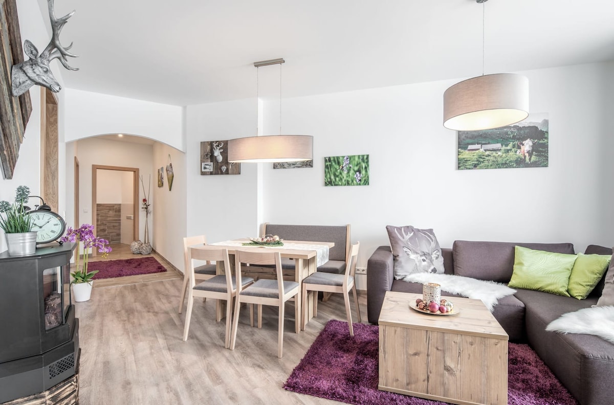 A bright living area features a cozy sofa, a wooden dining table with chairs, and modern light fixtures. Soft textures are provided by a rug on the floor, and artworks depicting nature adorn the walls, contributing to a tranquil environment.