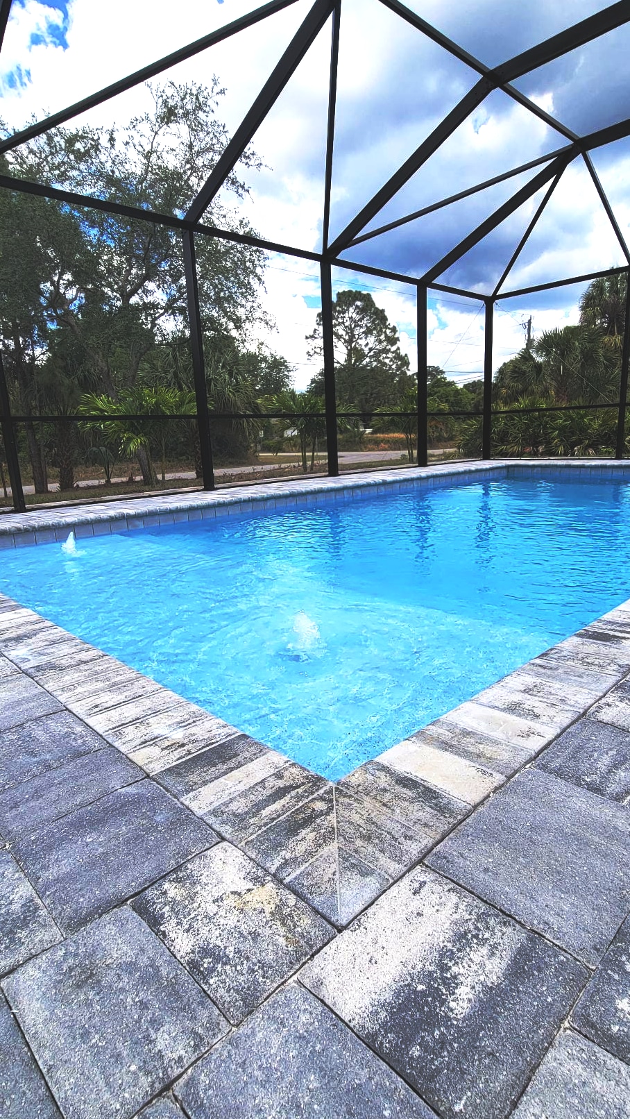 A heated pool is enclosed by a screened lanai, featuring clear blue water reflecting the sky. Stone pavers outline the pool area, while lush greenery is visible in the background, providing a serene outdoor setting.