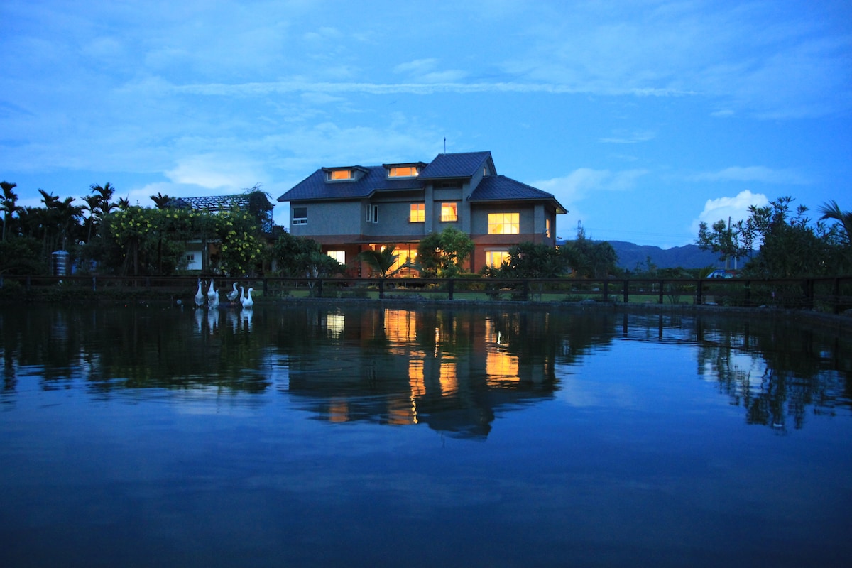 A serene lakeside view showcases a two-story villa, reflecting softly in the water. The home is framed by lush greenery, with strategically placed lights illuminating the windows. Three white ducks leisurely float on the calm water, contributing to a tranquil ambiance.