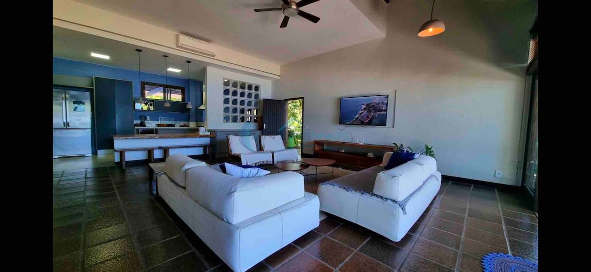 A spacious living area is highlighted by two large, light-colored couches facing a wooden coffee table. A ceiling fan circulates air above, and a blue accent wall enhances the modern design. Natural light filters in from large windows, offering views of the outdoors.