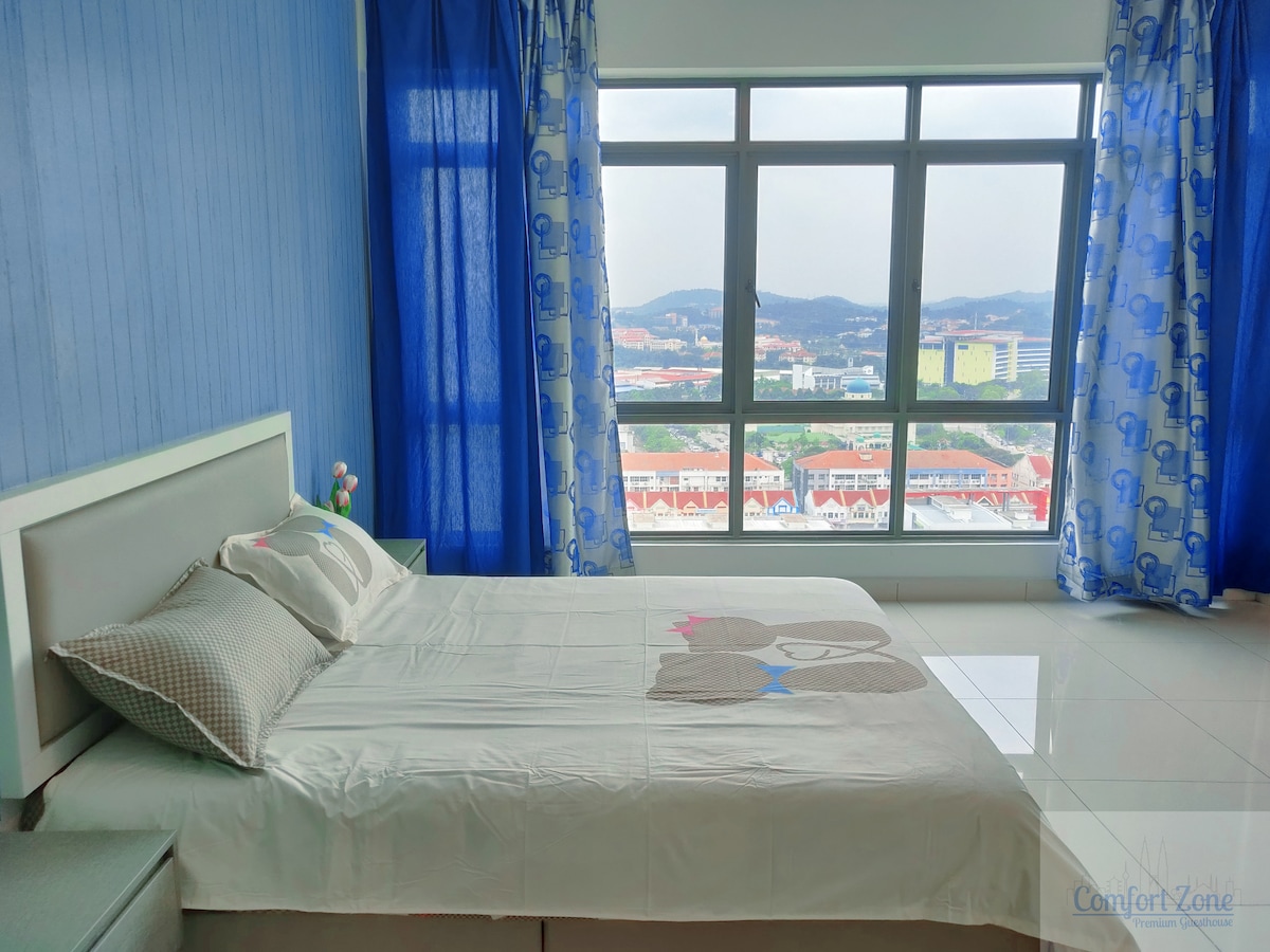 A bright and airy studio unit showcases a spacious bed adorned with a playful design. Large windows framed with blue curtains provide a view of the surroundings, allowing natural light to fill the room. A small decorative item is placed on the bedside table.