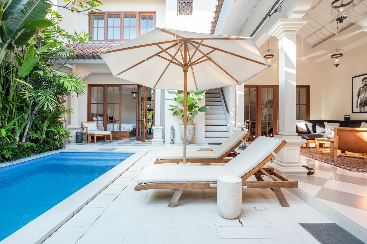 A sunlit outdoor area features two wooden lounge chairs positioned beside a clear blue pool. An umbrella provides shade, and lush greenery surrounds the space. The villa's elegant architecture is highlighted by wooden doors and bright interior spaces visible in the background.